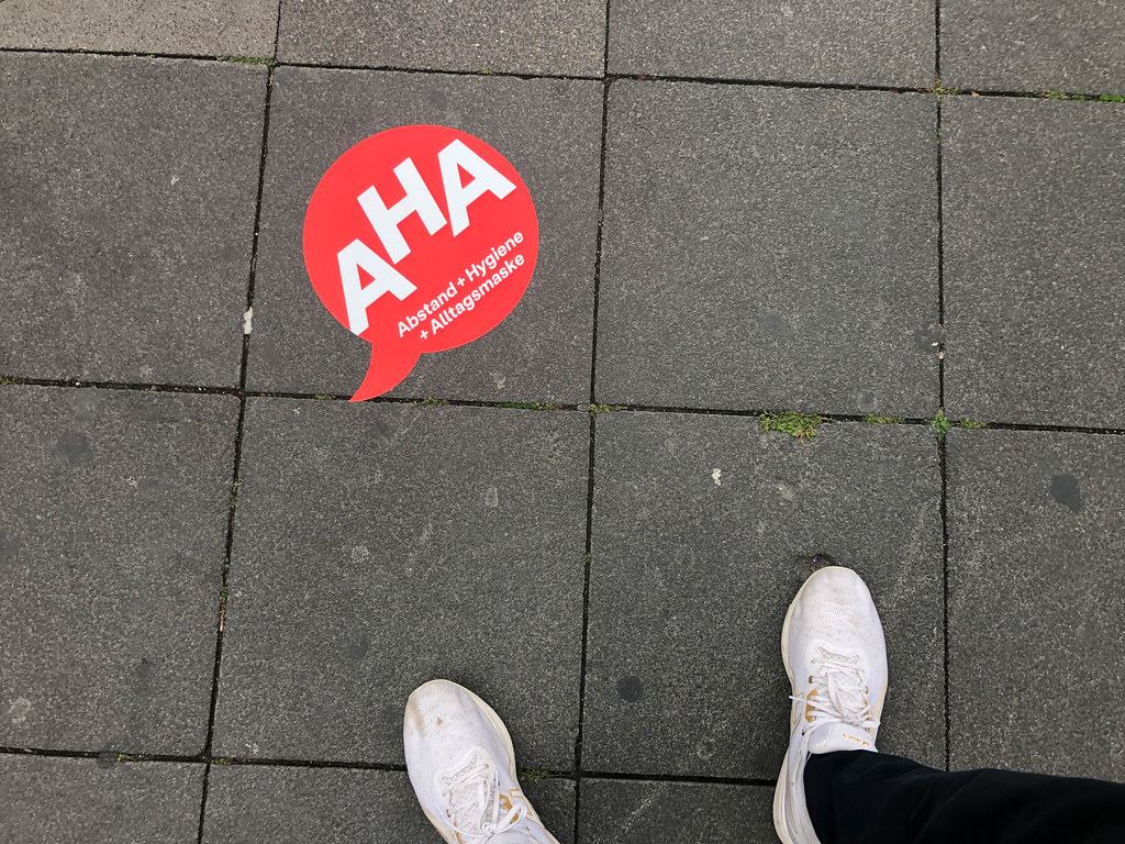 Füße und Sticker auf der Straße mit der AHA-Formel für den Corona-Alltag (Abstand, Hygiene, Alltagsmaske)