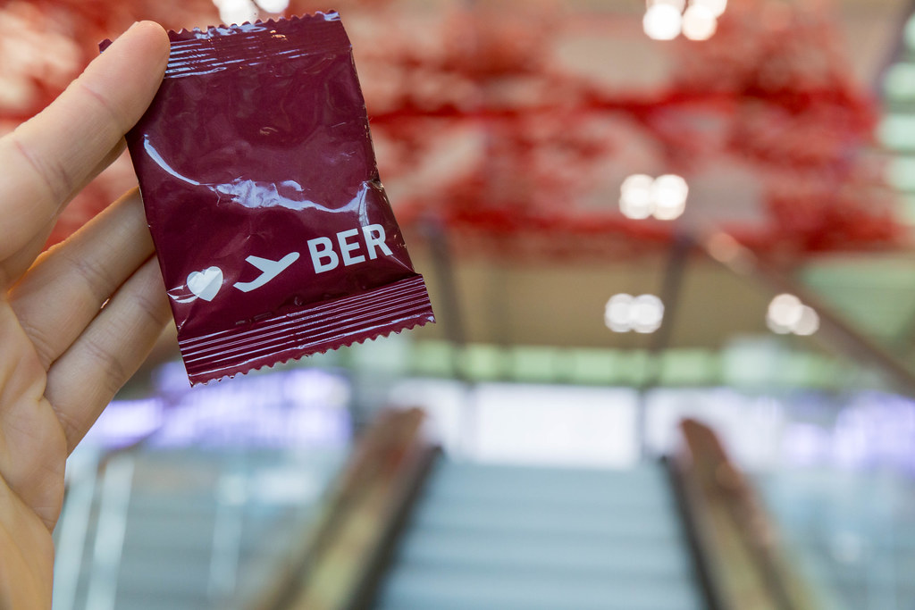 Hand mit rotem Gadget des Flughafens BER vor leeren Rolltreppen und Pae Whites rotem Kunstwerk