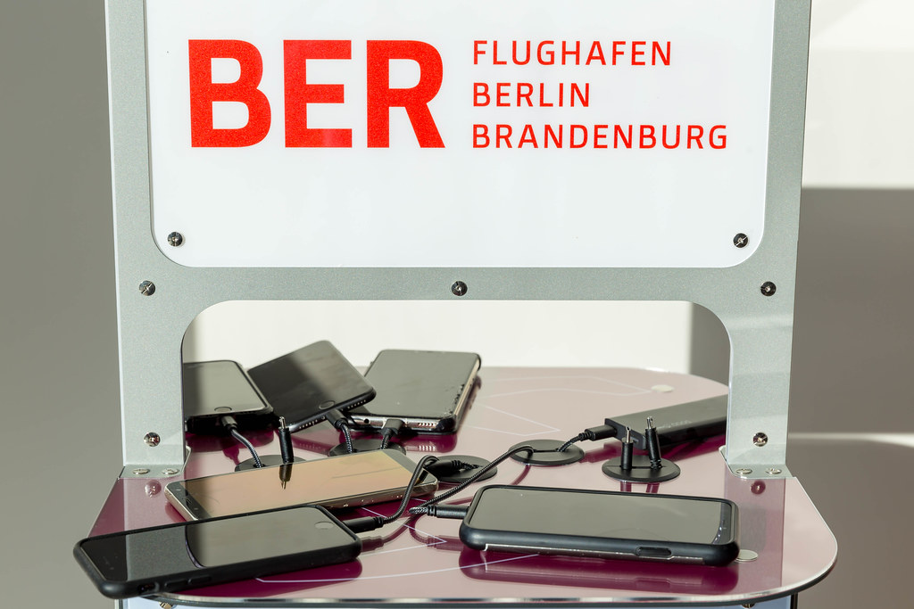 Sechs Handys und eine Powerbank werden an einer Ladestation mit BER-Flughafen-Logo aufgeladen