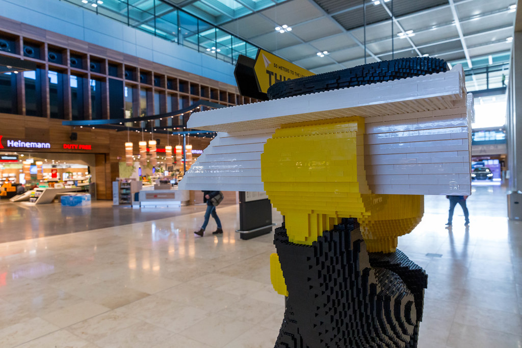 Nahaufnahme: Papierflugzeug aus Legosteine in der Hand einer lebensgroßen Piloten-Figur am Flughafen BER