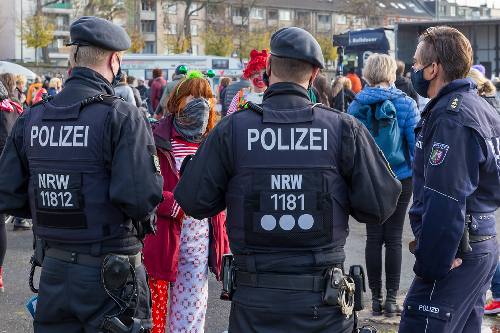 Polizisten reden mit einer Frau mit Halstuch als Mundschutz, in traditionellen Farben von Köln verkleidet