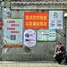 A man crouches by a rubbish collection station checking his smartphone to deal with business, and the wall behind him is covered with a variety of fantastic signs.